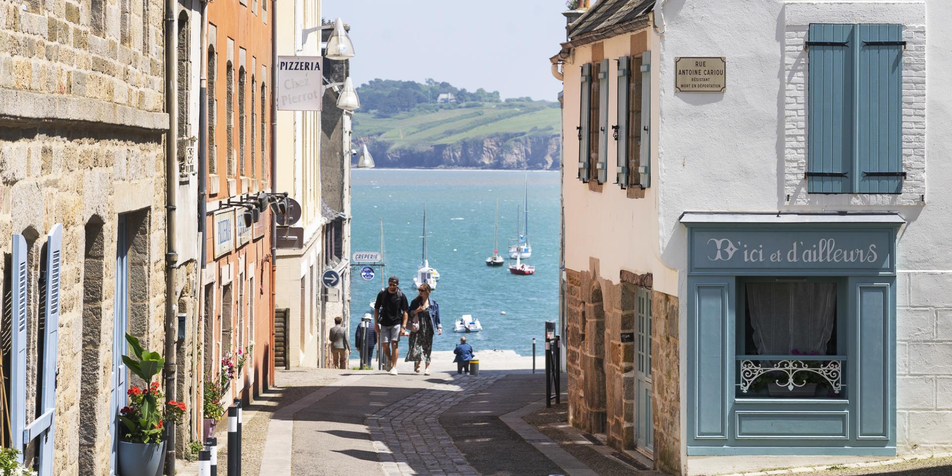 Un couple remonte la Rue depuis le port du Romseur. De part et d'autre, il ya des maison à 2 ou 3 étages. Derrière on voit la Baie de Douarnenez avec une belle météo