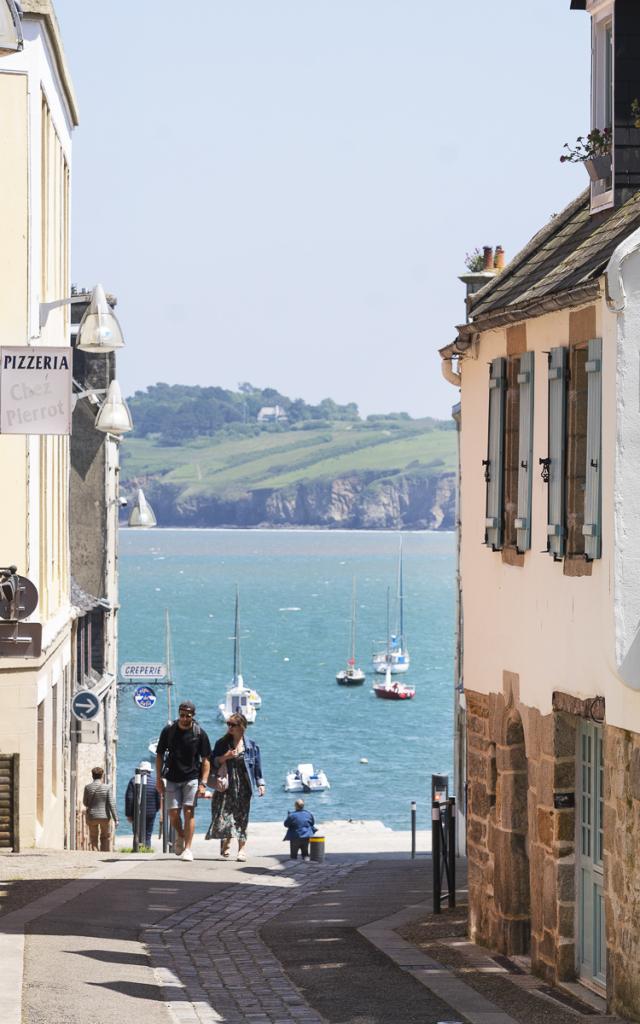 Un couple remonte la Rue depuis le port du Romseur. De part et d'autre, il ya des maison à 2 ou 3 étages. Derrière on voit la Baie de Douarnenez avec une belle météo
