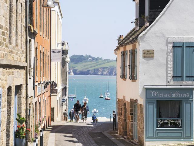 Un couple remonte la Rue depuis le port du Romseur. De part et d'autre, il ya des maison à 2 ou 3 étages. Derrière on voit la Baie de Douarnenez avec une belle météo