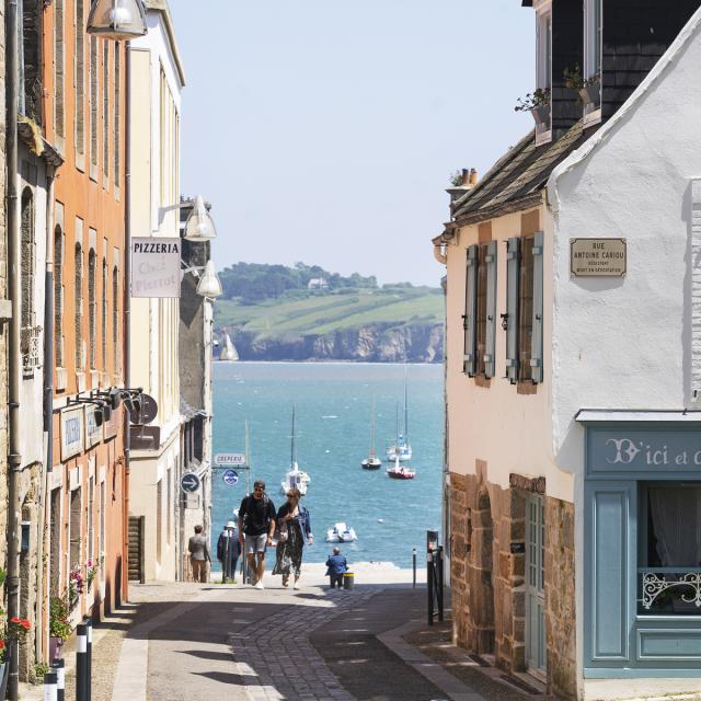Un couple remonte la Rue depuis le port du Romseur. De part et d'autre, il ya des maison à 2 ou 3 étages. Derrière on voit la Baie de Douarnenez avec une belle météo